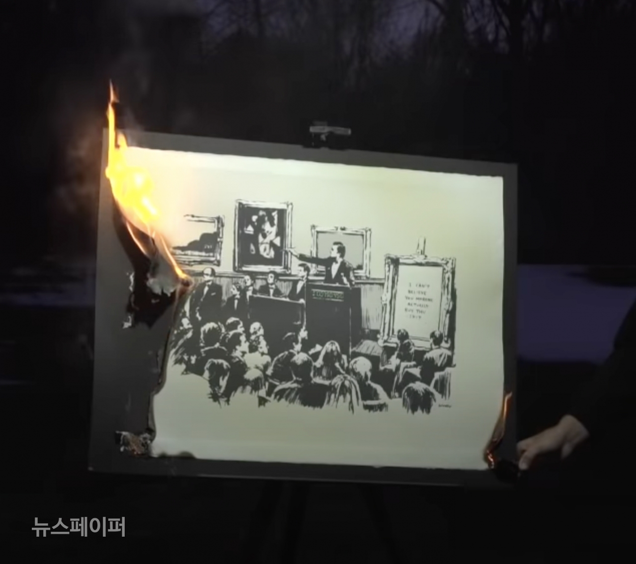 [BurntBanksy의 유튜브 채널의 한 장면. 뱅크시의 판화가 불에 타고 있다. 오른쪽에는 라이터를 들고 불을 붙이는 손.]